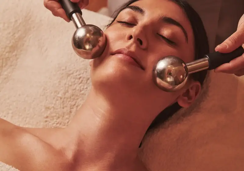 Person receiving a facial massage with two metal massage globes while lying wrapped in a soft towel.