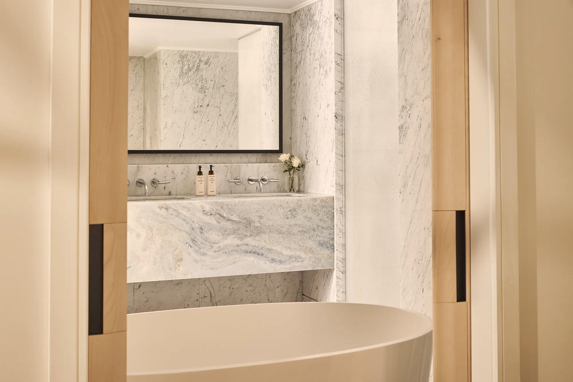 Freestanding bathtub before marble vanity and mirror in bright bathroom with pale wood doors and stone walls.