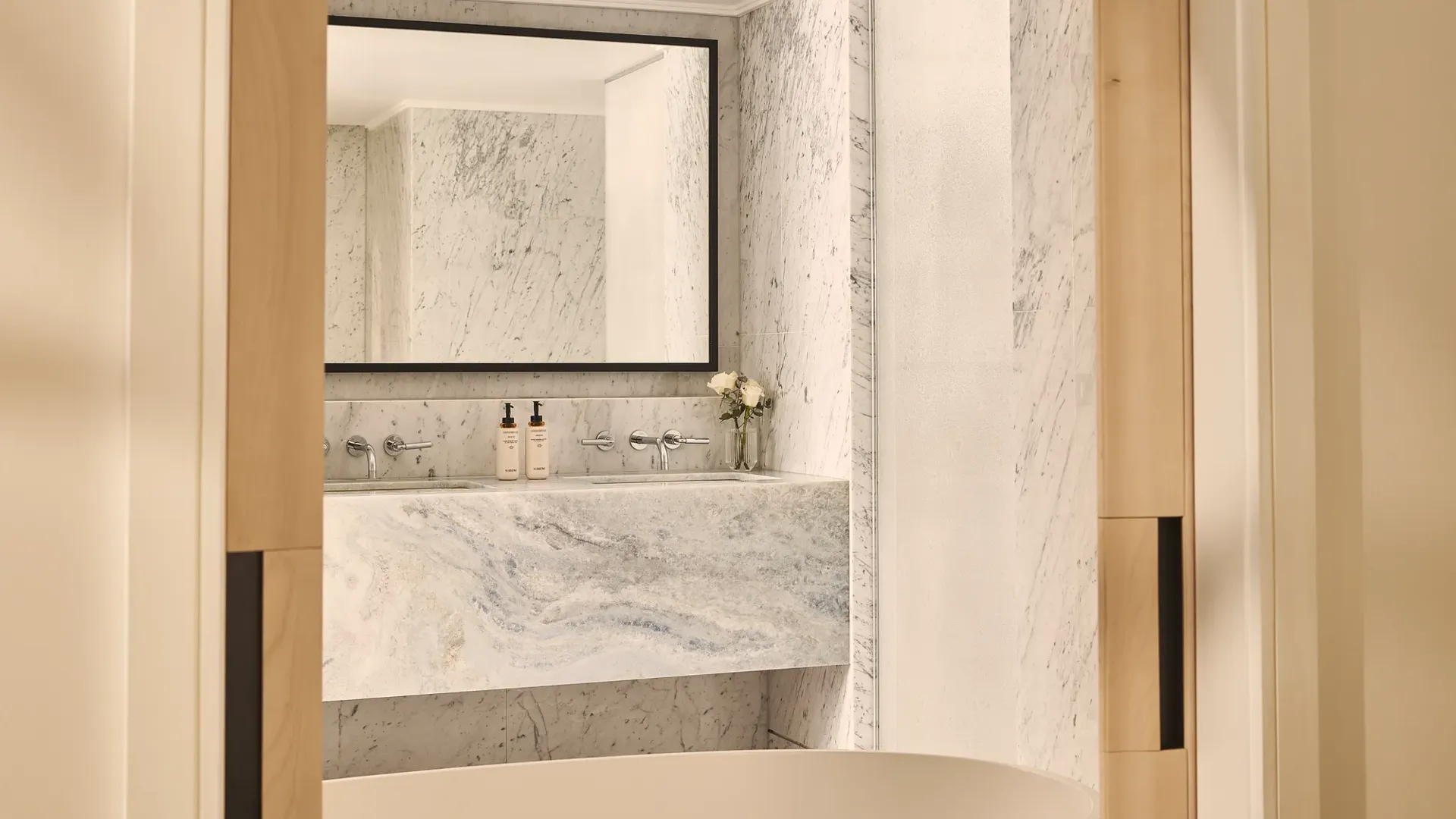 Freestanding bathtub before marble vanity and mirror in bright bathroom with pale wood doors and stone walls.