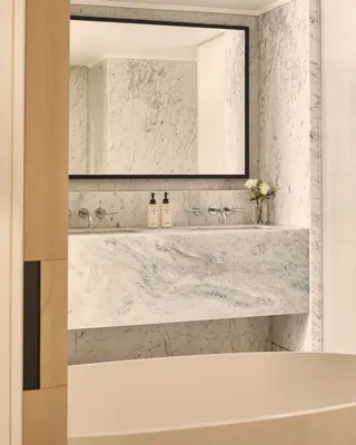 Freestanding bathtub before marble vanity and mirror in bright bathroom with pale wood doors and stone walls.