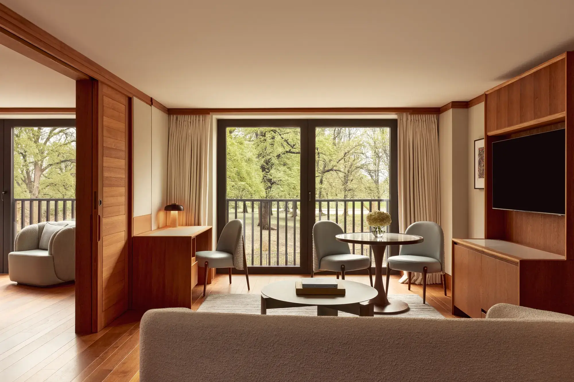 Contemporary suite living space with pale wood floors, a desk, round dining table with blue chairs, and large windows opening onto views of Hyde Park.