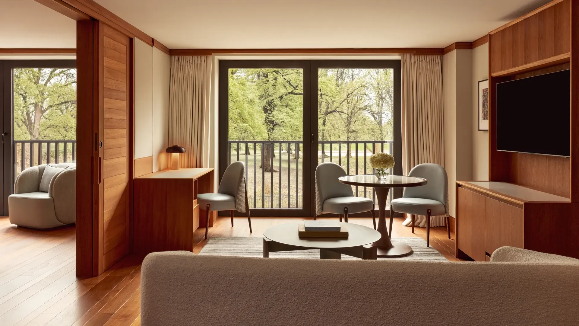 Contemporary suite living space with pale wood floors, a desk, round dining table with blue chairs, and large windows opening onto views of Hyde Park.