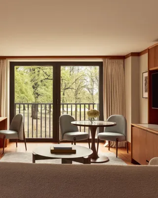 Contemporary suite living space with pale wood floors, a desk, round dining table with blue chairs, and large windows opening onto views of Hyde Park.