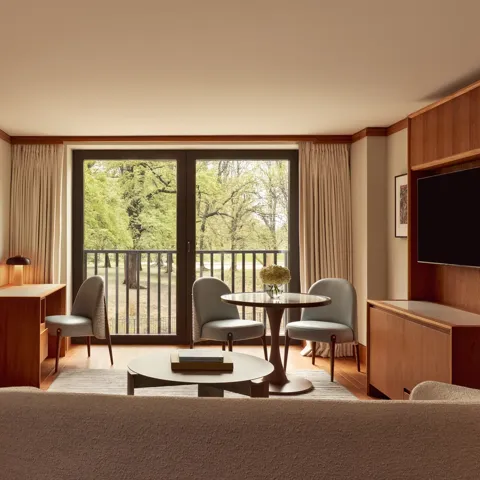 Contemporary suite living space with pale wood floors, a desk, round dining table with blue chairs, and large windows opening onto views of Hyde Park.