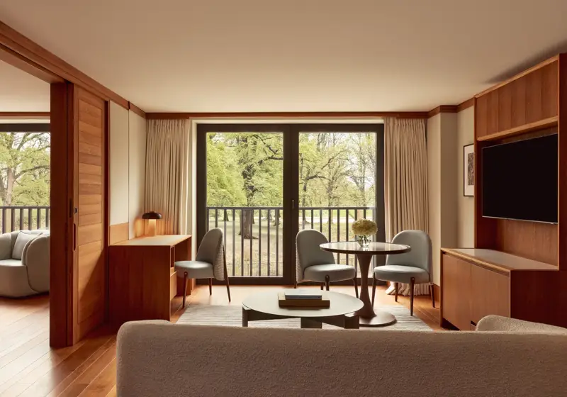 Contemporary suite living space with pale wood floors, a desk, round dining table with blue chairs, and large windows opening onto views of Hyde Park.