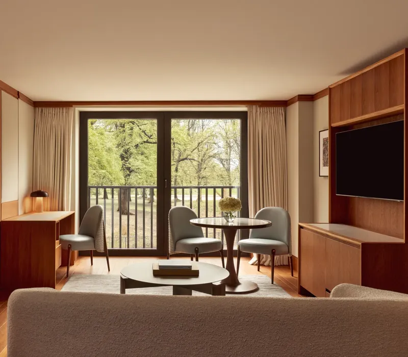 Contemporary suite living space with pale wood floors, a desk, round dining table with blue chairs, and large windows opening onto views of Hyde Park.
