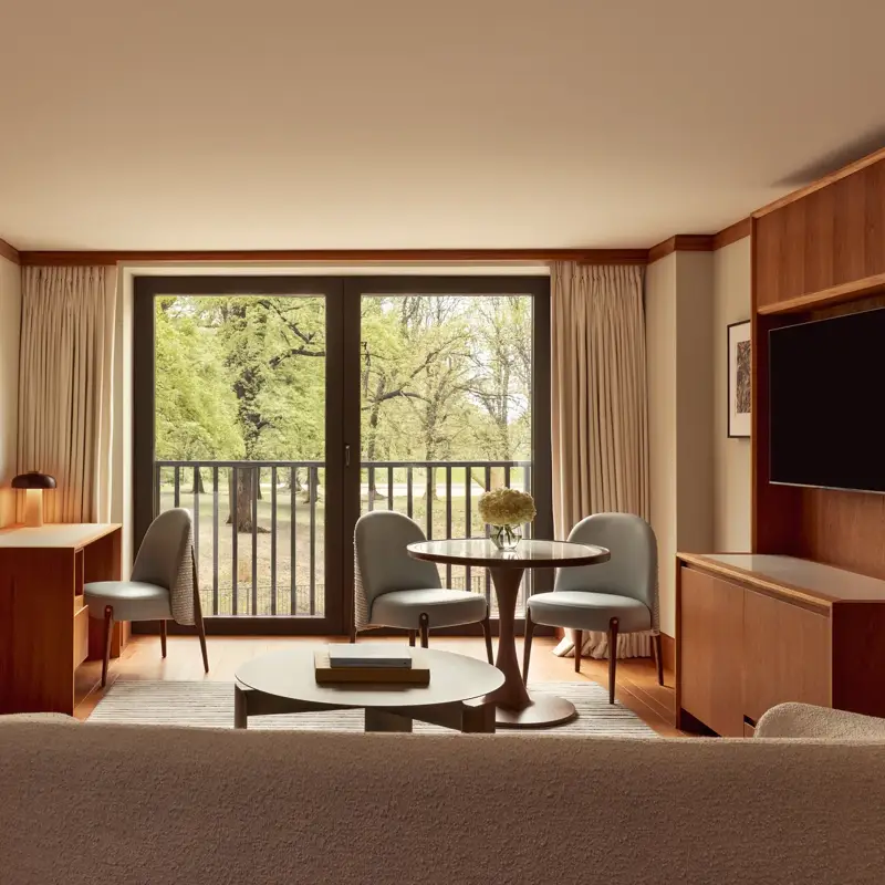 Contemporary suite living space with pale wood floors, a desk, round dining table with blue chairs, and large windows opening onto views of Hyde Park.