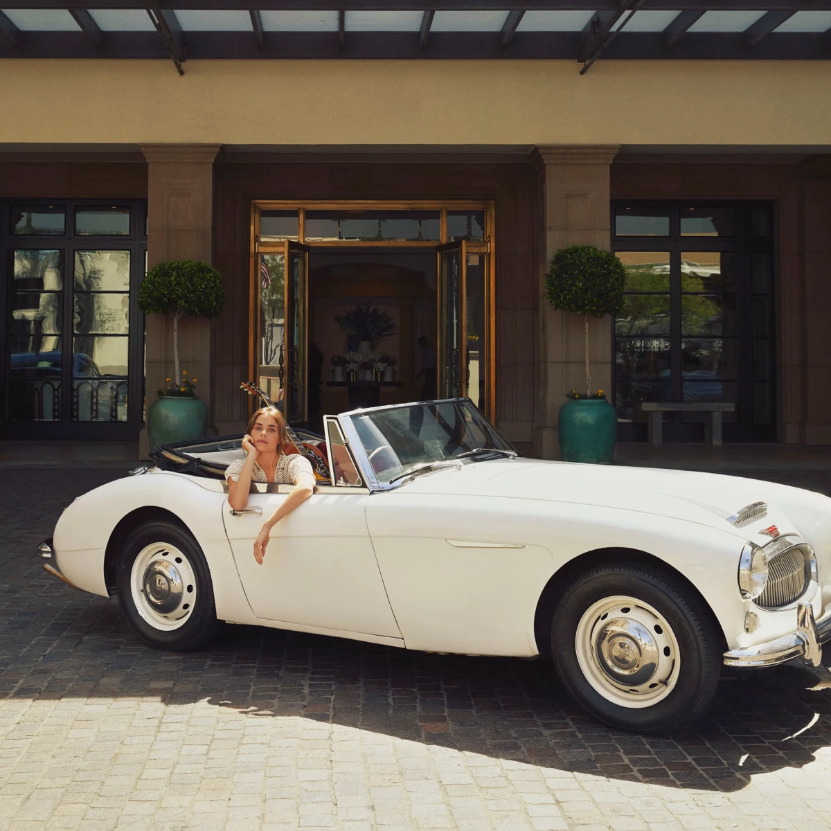 Guest seated in a white vintage convertible outside the hotel entrance, framed by glass doors, planters, and a covered portico.