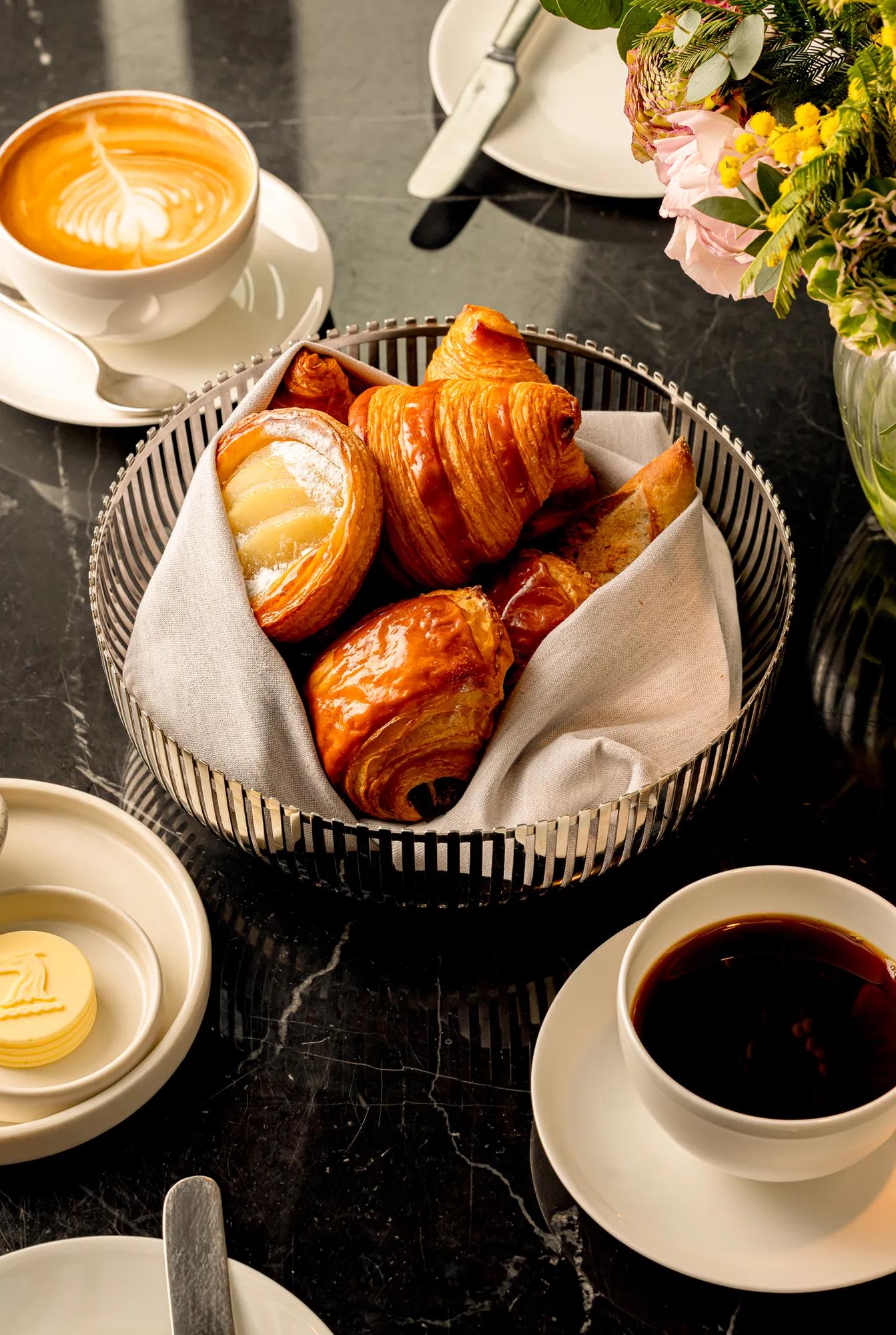 Basket of pastries on a marble table, served with coffee, a latte, and butter.