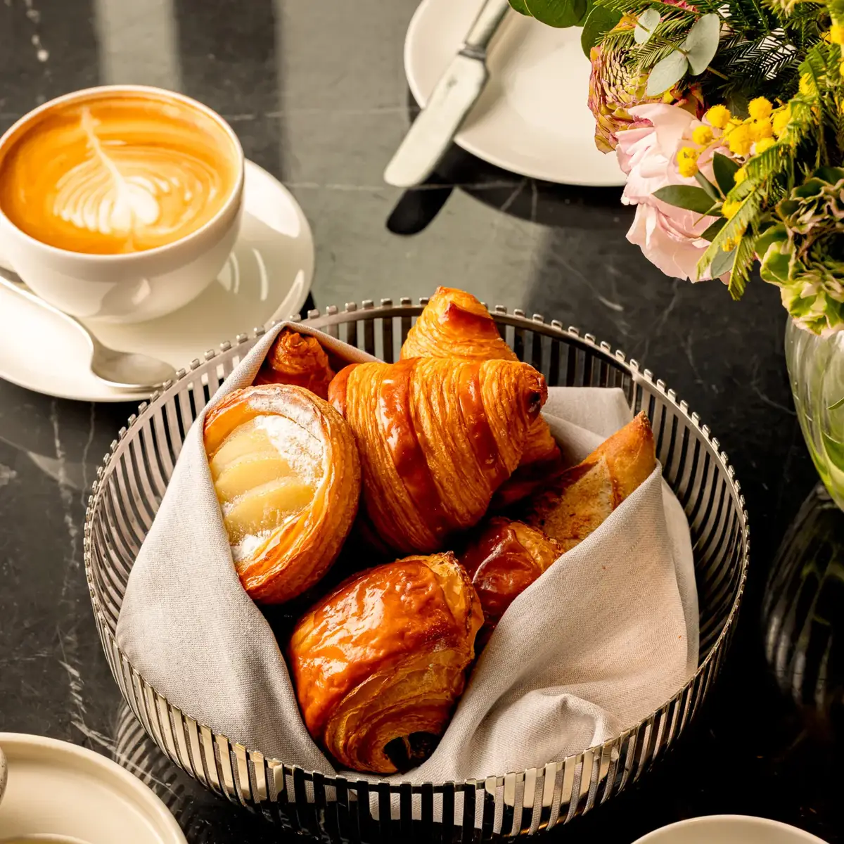 Basket of pastries on a marble table, served with coffee, a latte, and butter.