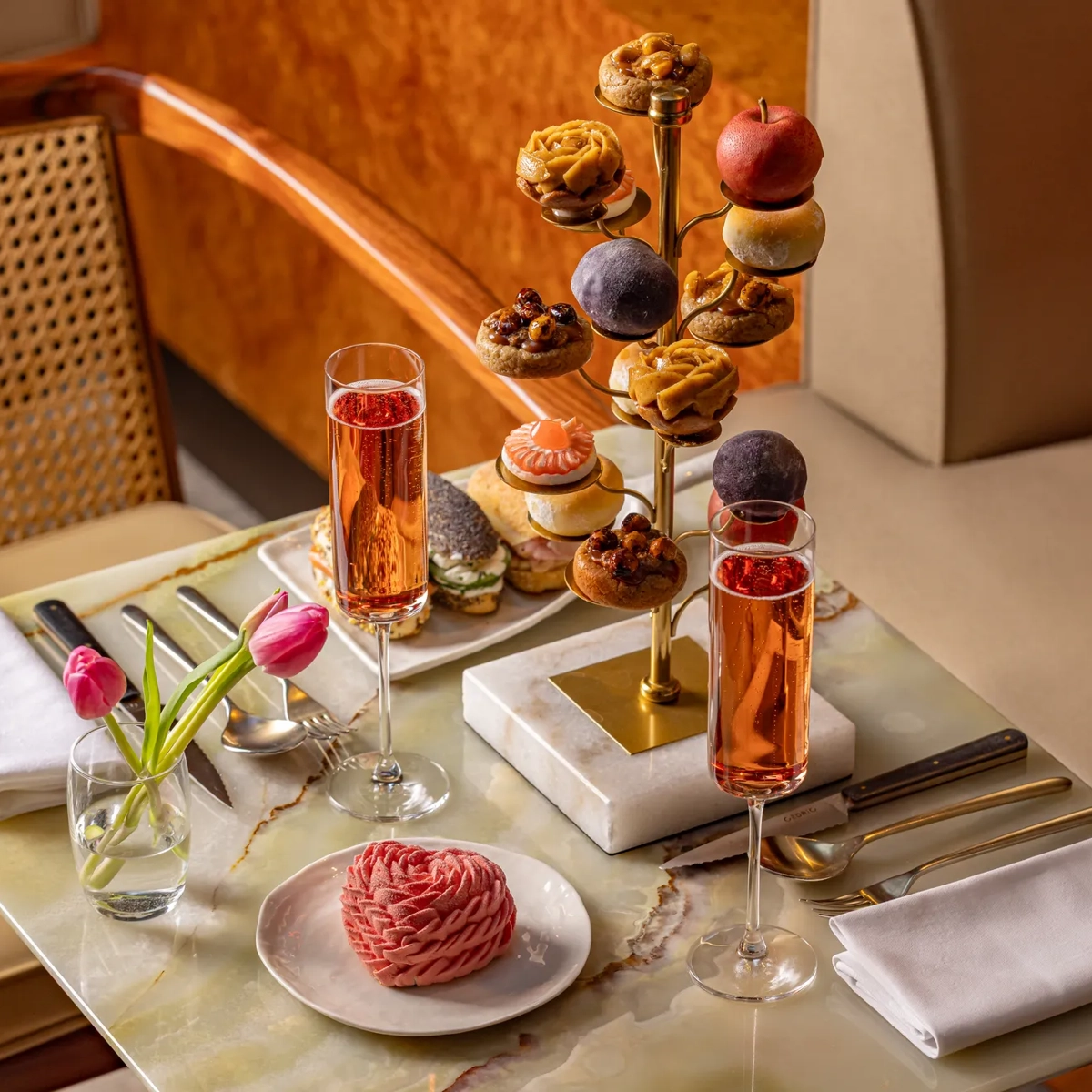 Afternoon tea with pastries on a tiered stand, champagne flutes, pink dessert, tulips, and neatly set table in an elegant lounge.
