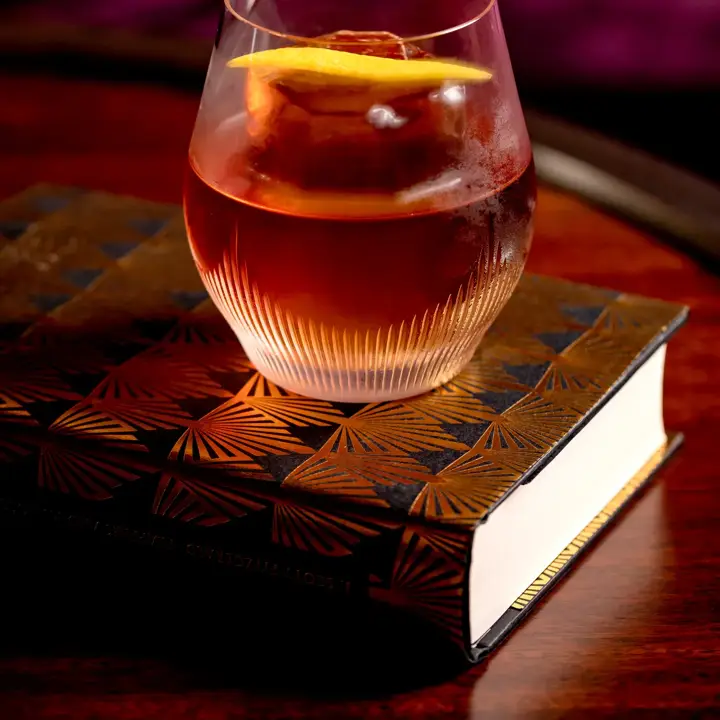 Amber cocktail with citrus garnish served in a cut-glass tumbler on a decorative book