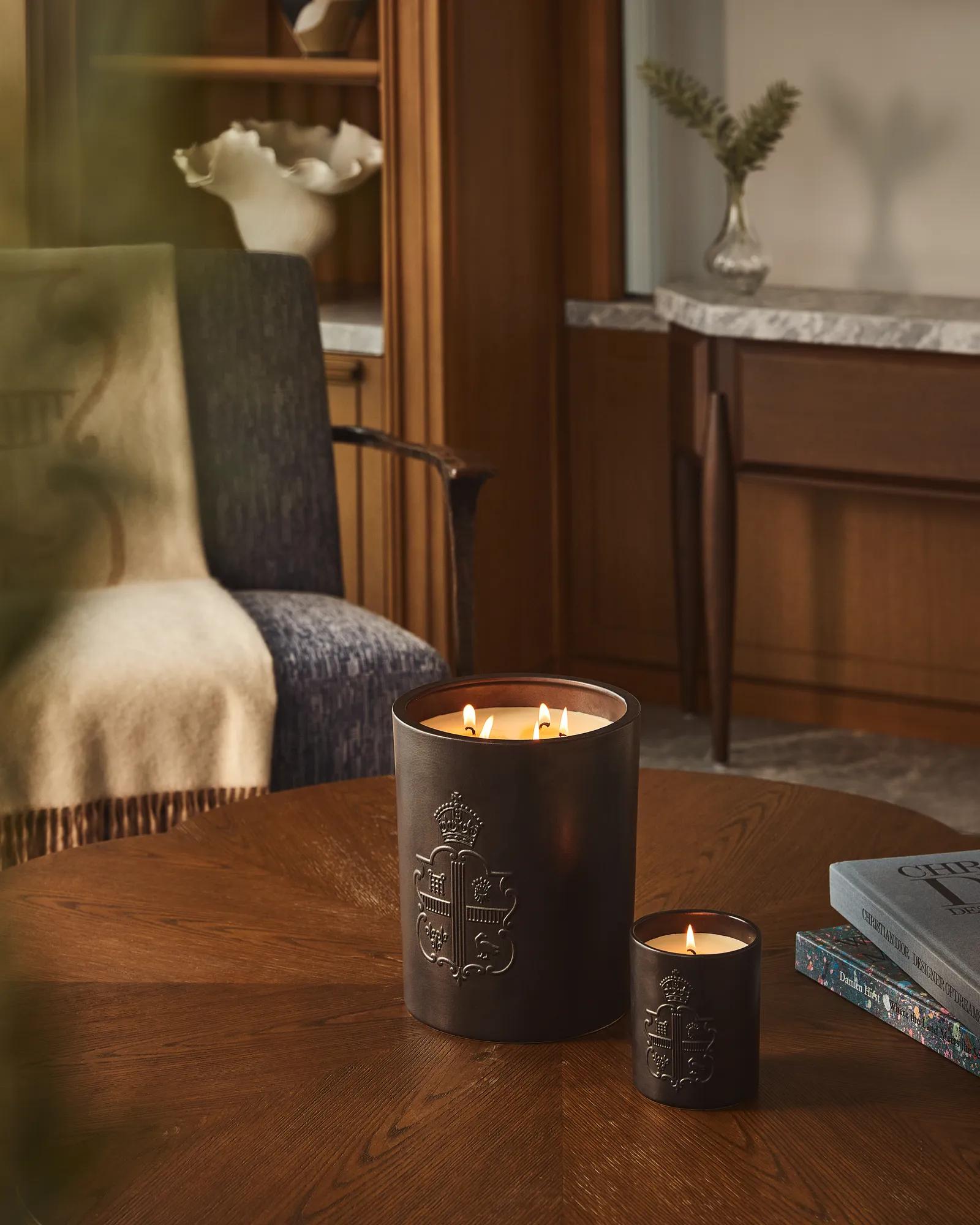Two lit candles with embossed crest on wooden table beside books, with armchair and wood-panelled interior behind.
 