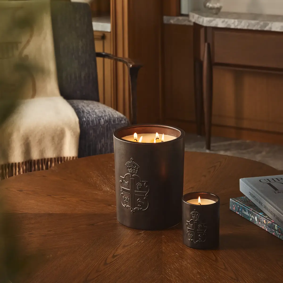 Two lit candles with embossed crest on wooden table beside books, with armchair and wood-panelled interior behind.
 