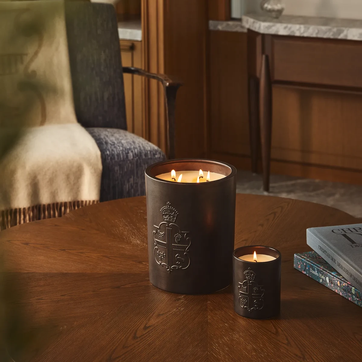 Two lit candles with embossed crest on wooden table beside books, with armchair and wood-panelled interior behind.
 