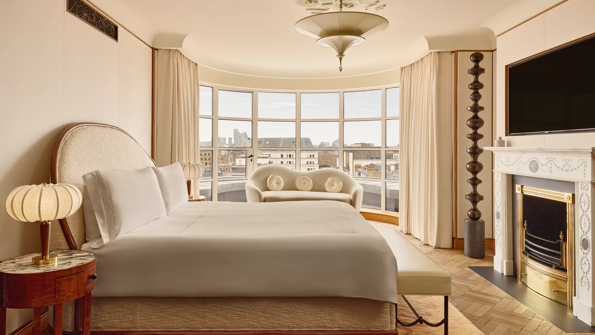Spacious bedroom with a curved window overlooking London, featuring a king-size bed, cream sofa, marble fireplace, and warm neutral tones with Art Deco-inspired details.