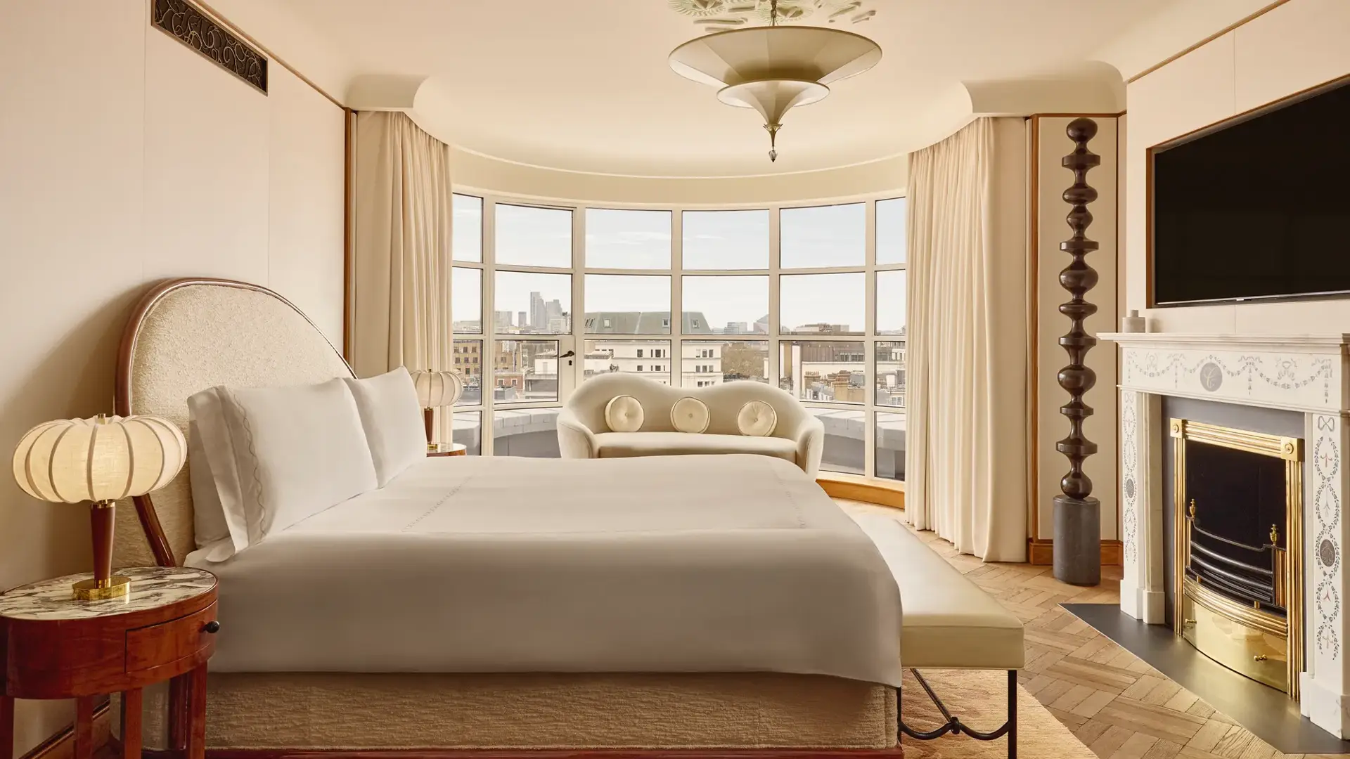 Spacious bedroom with a curved window overlooking London, featuring a king-size bed, cream sofa, marble fireplace, and warm neutral tones with Art Deco-inspired details.