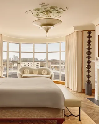 Spacious bedroom with a curved window overlooking London, featuring a king-size bed, cream sofa, marble fireplace, and warm neutral tones with Art Deco-inspired details.