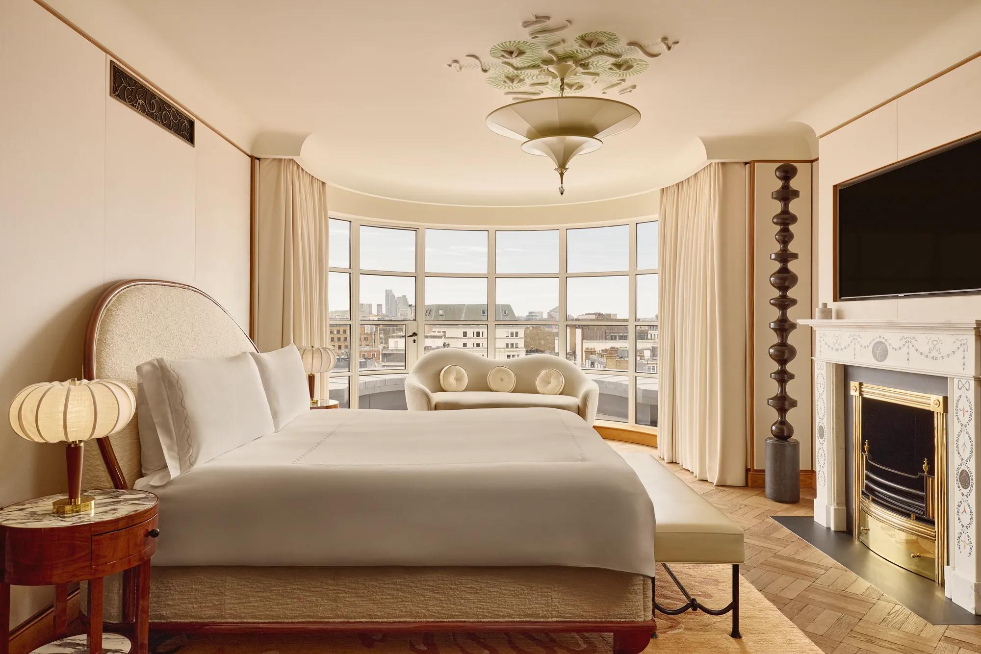 Spacious bedroom with a curved window overlooking London, featuring a king-size bed, cream sofa, marble fireplace, and warm neutral tones with Art Deco-inspired details.