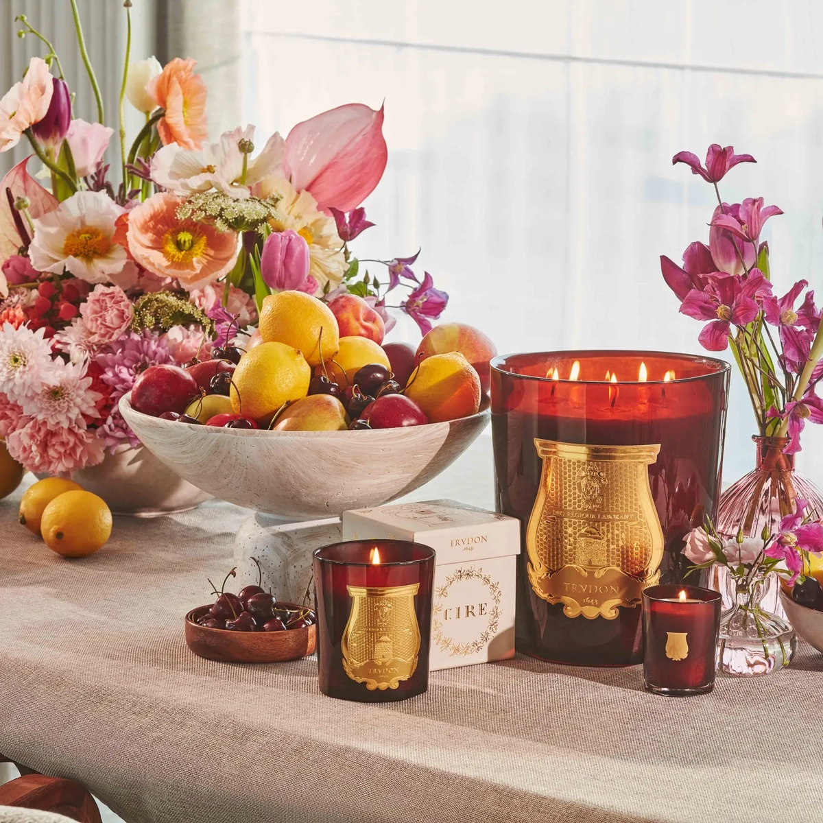 Still life of lit candles, bowls of fruit and pink flowers arranged on a linen-covered table by a window.