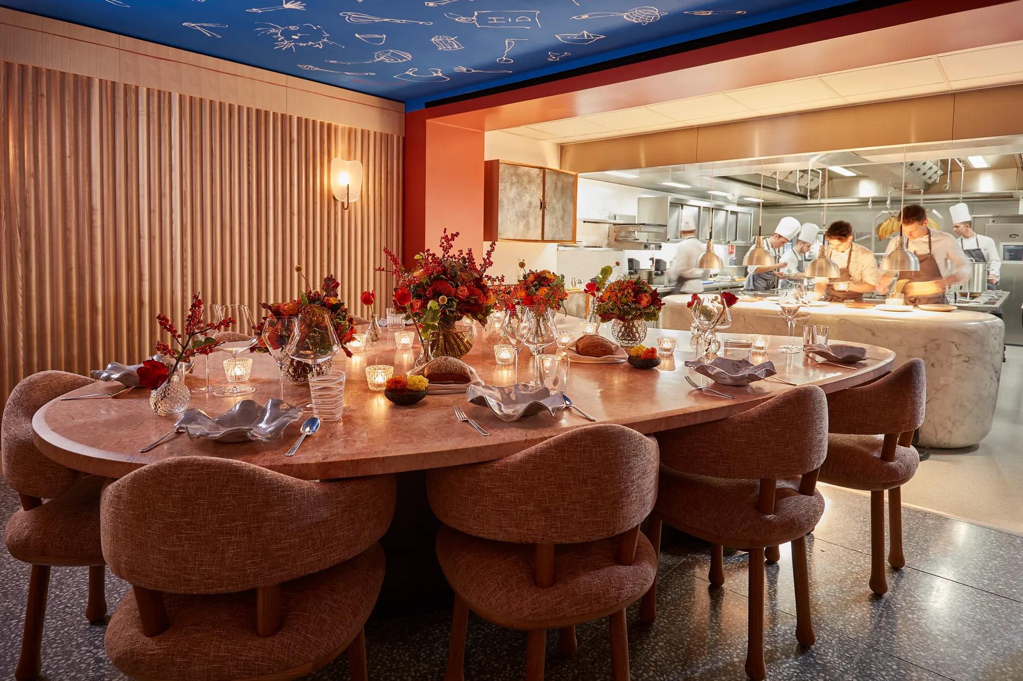 Private dining table set with candles and flowers beside an open kitchen, where chefs prepare dishes behind a glass partition.