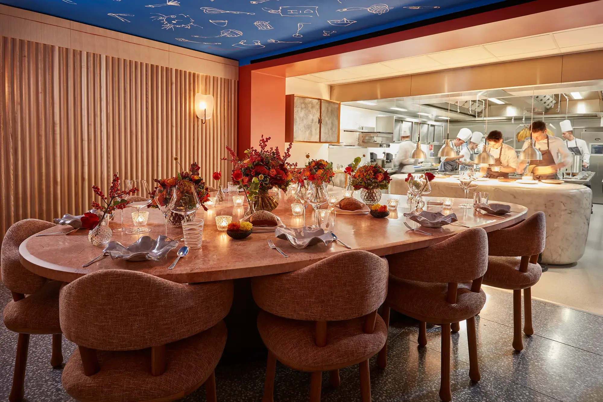Private dining table set with candles and flowers beside an open kitchen, where chefs prepare dishes behind a glass partition.