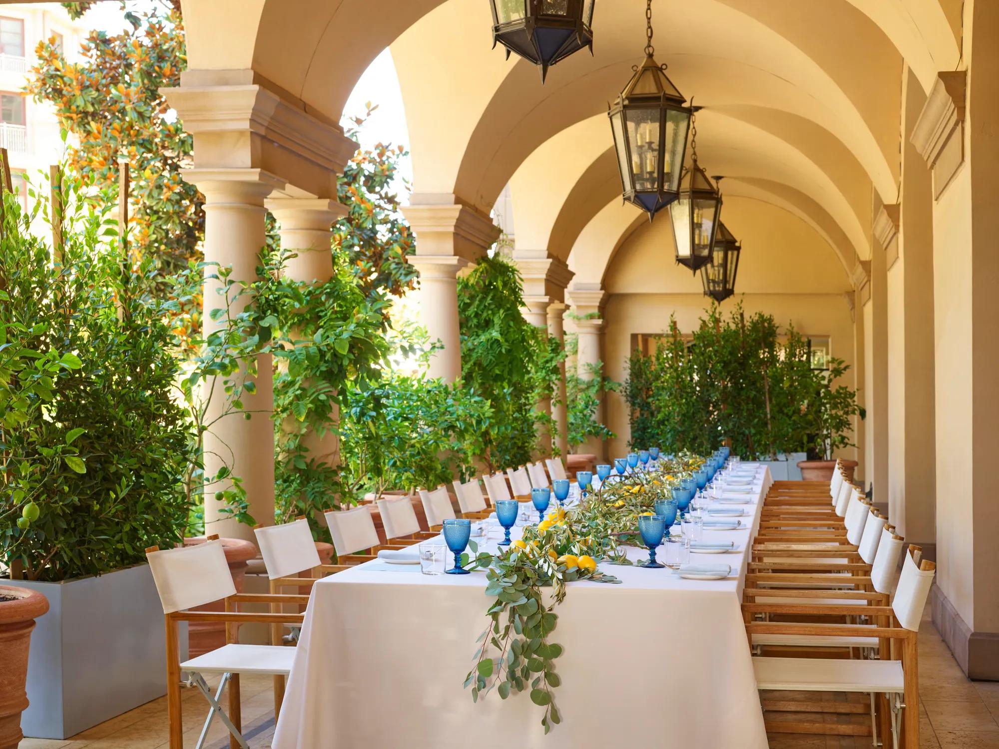 Arched outdoor corridor set for a luncheon; twin rows of chairs flank a white-clothed table with blue goblets and citrus greenery.