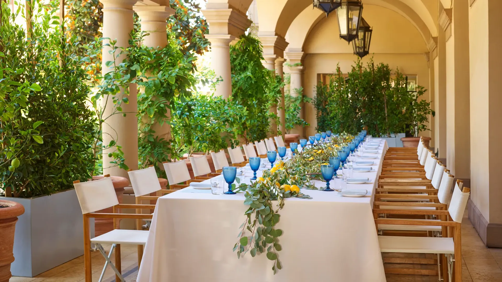 Arched outdoor corridor set for a luncheon; twin rows of chairs flank a white-clothed table with blue goblets and citrus greenery.