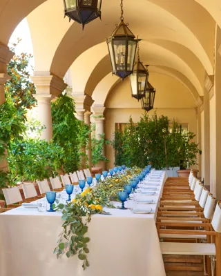 Arched outdoor corridor set for a luncheon; twin rows of chairs flank a white-clothed table with blue goblets and citrus greenery.