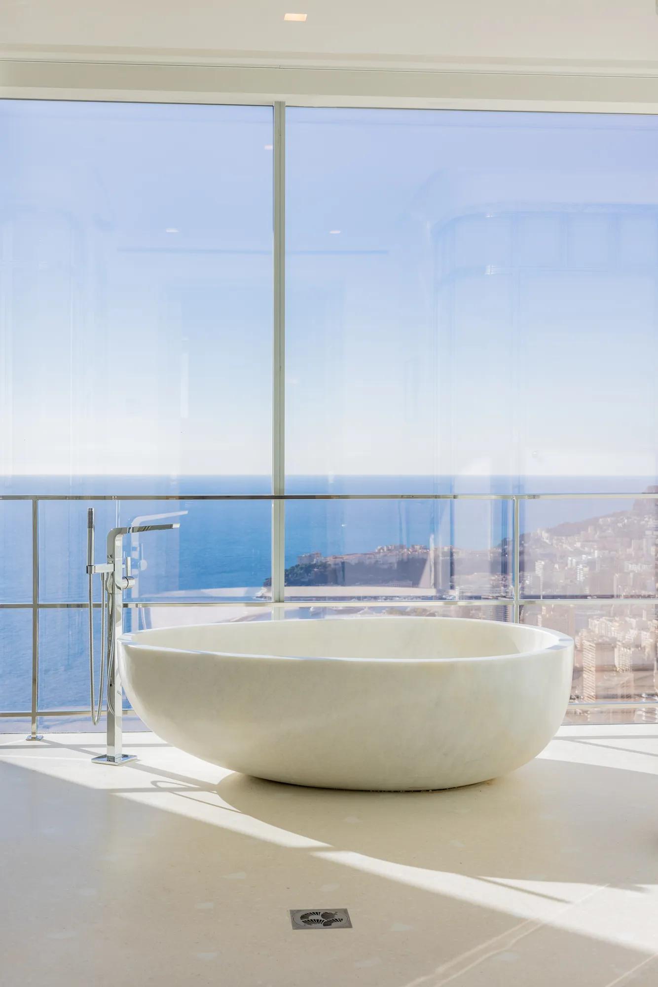 Baignoire blanche autoportante devant de larges baies vitrées, offrant une vue panoramique sur la mer et la ville côtière.