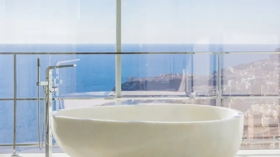 Baignoire blanche autoportante devant de larges baies vitrées, offrant une vue panoramique sur la mer et la ville côtière.