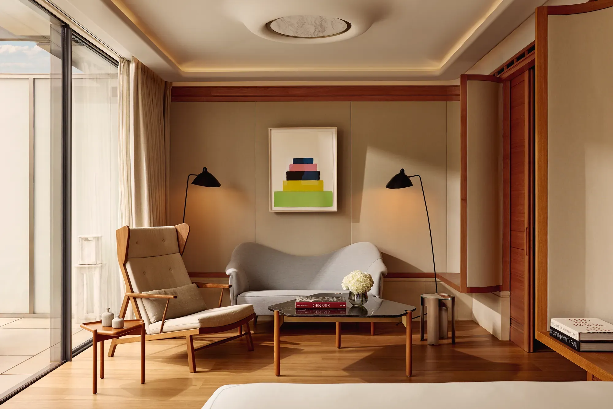 A bright suite seating area with a tall cream armchair, a small sofa, and a glass coffee table topped with flowers and books, set beside floor-to-ceiling windows leading to the terrace.