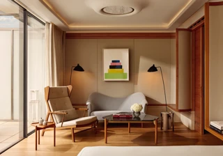 A bright suite seating area with a tall cream armchair, a small sofa, and a glass coffee table topped with flowers and books, set beside floor-to-ceiling windows leading to the terrace.