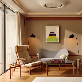 A bright suite seating area with a tall cream armchair, a small sofa, and a glass coffee table topped with flowers and books, set beside floor-to-ceiling windows leading to the terrace.