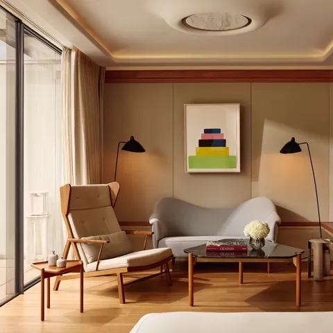 A bright suite seating area with a tall cream armchair, a small sofa, and a glass coffee table topped with flowers and books, set beside floor-to-ceiling windows leading to the terrace.