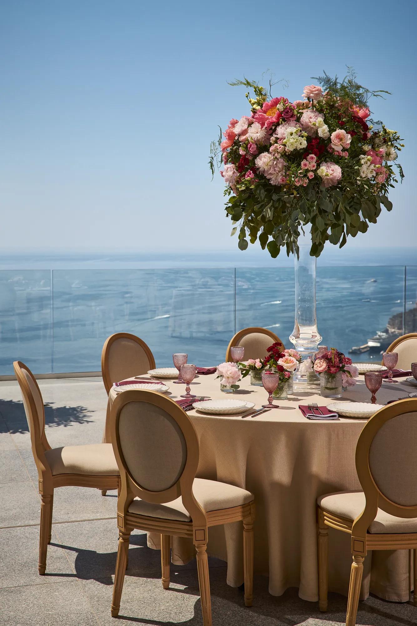 Table ronde élégante dressée pour un mariage, avec des chaises beiges, de la verrerie rose et une grande composition florale de roses et d’hortensias, donnant sur la mer.