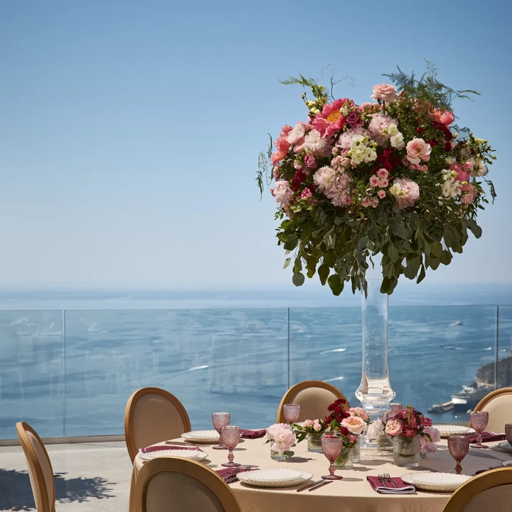 Table ronde élégante dressée pour un mariage, avec des chaises beiges, de la verrerie rose et une grande composition florale de roses et d’hortensias, donnant sur la mer.