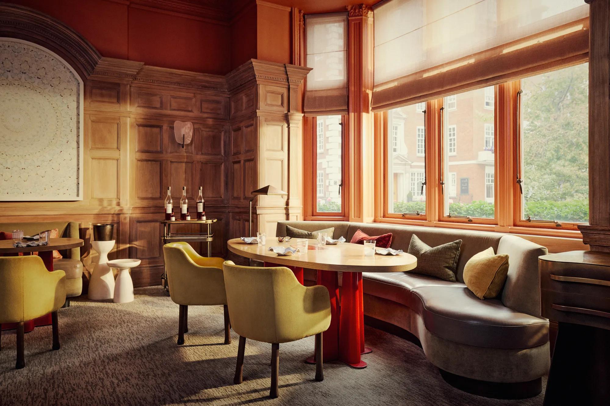 Elegant dining room with wood-panelled walls, curved banquette seating, and sunlit windows overlooking a tree-lined street.
