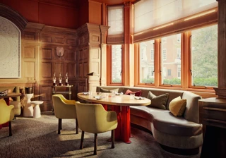 Elegant dining room with wood-panelled walls, curved banquette seating, and sunlit windows overlooking a tree-lined street.