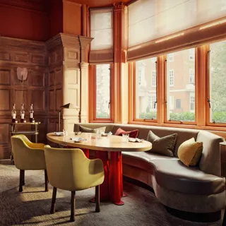 Elegant dining room with wood-panelled walls, curved banquette seating, and sunlit windows overlooking a tree-lined street.