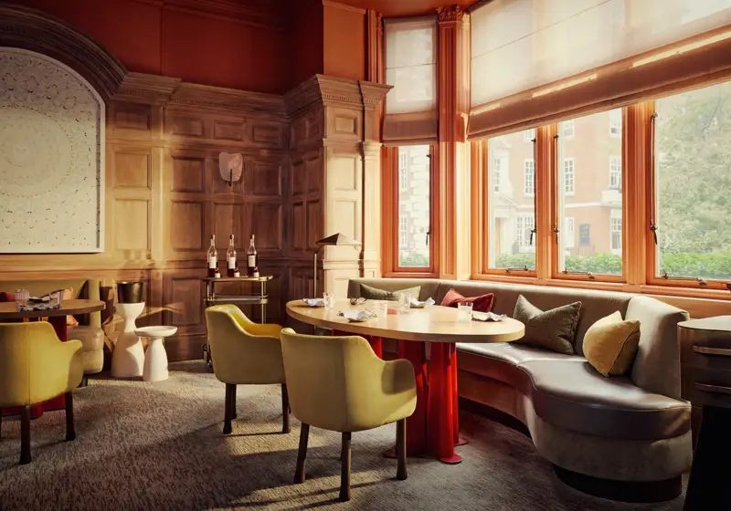 Elegant dining room with wood-panelled walls, curved banquette seating, and sunlit windows overlooking a tree-lined street.