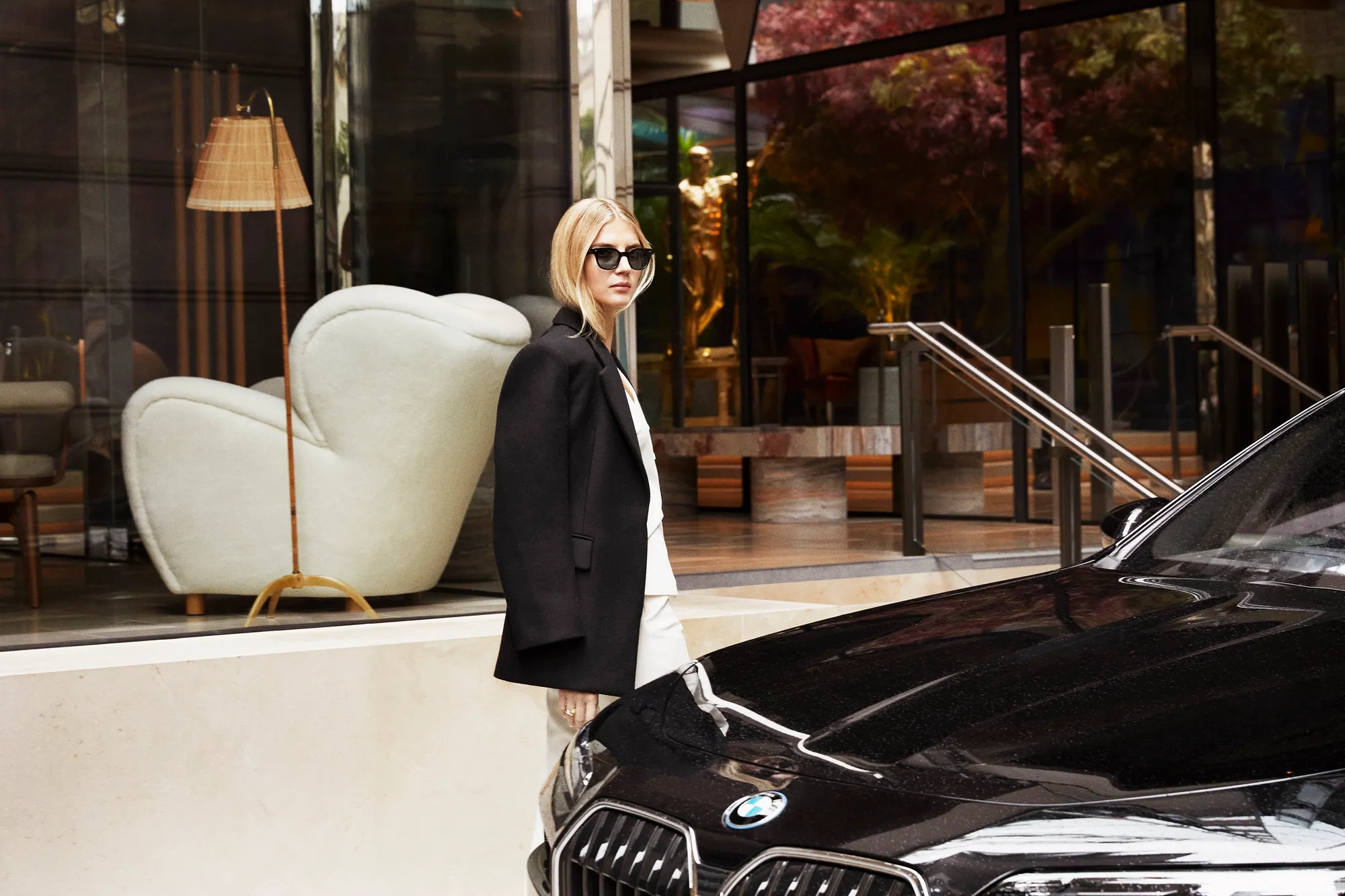 Stylish woman in sunglasses and black blazer stands beside a black BMW outside a modern glass-fronted building with elegant seating inside.