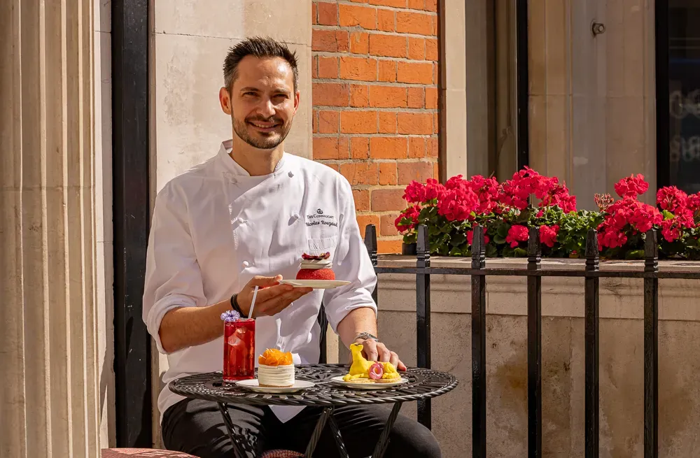 Chef seated outdoors at café table holding dessert, with pastries and drinks in sunlight.