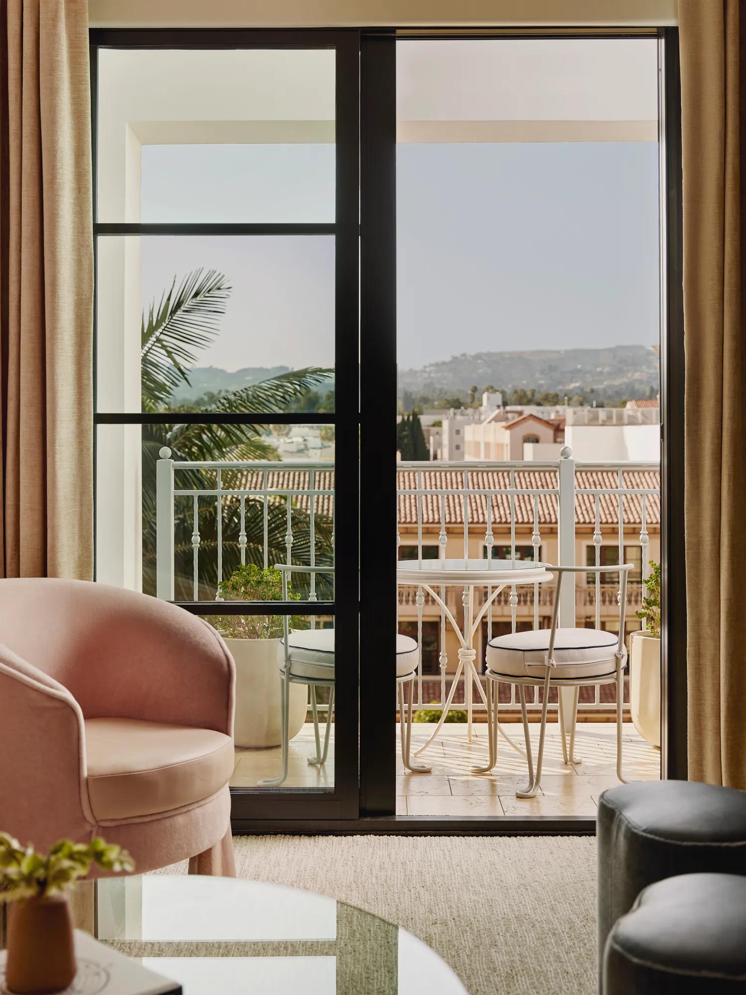 View through sliding doors to a balcony with a round table and two chairs, overlooking tiled rooftops and distant hills; lounge seating in foreground.
