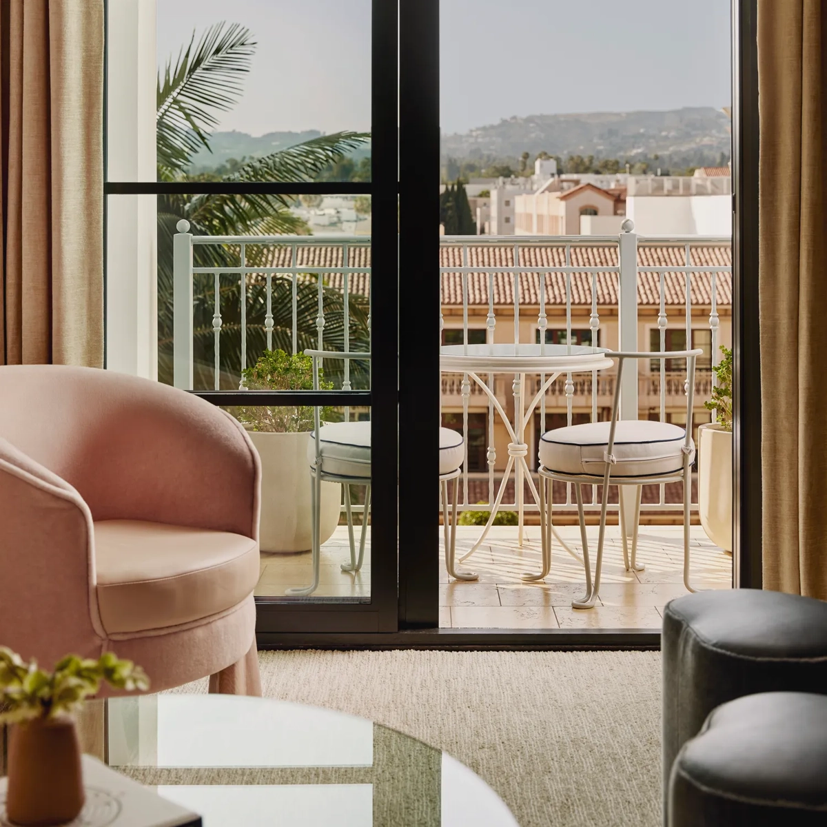 View through sliding doors to a balcony with a round table and two chairs, overlooking tiled rooftops and distant hills; lounge seating in foreground.