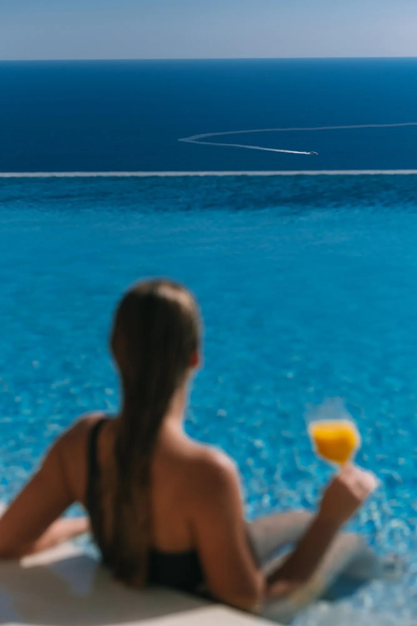 Personne se relaxant dans une piscine à débordement avec un verre, regardant la mer et le sillage d’un bateau.