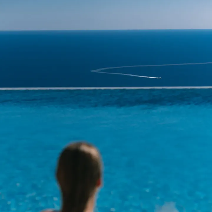Personne se relaxant dans une piscine à débordement avec un verre, regardant la mer et le sillage d’un bateau.