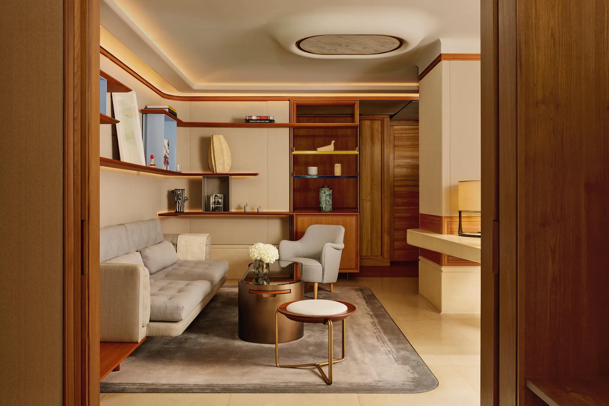 A stylish sitting room with a pale grey sofa, armchair, and round coffee table, framed by wooden shelving displaying books and decorative pieces