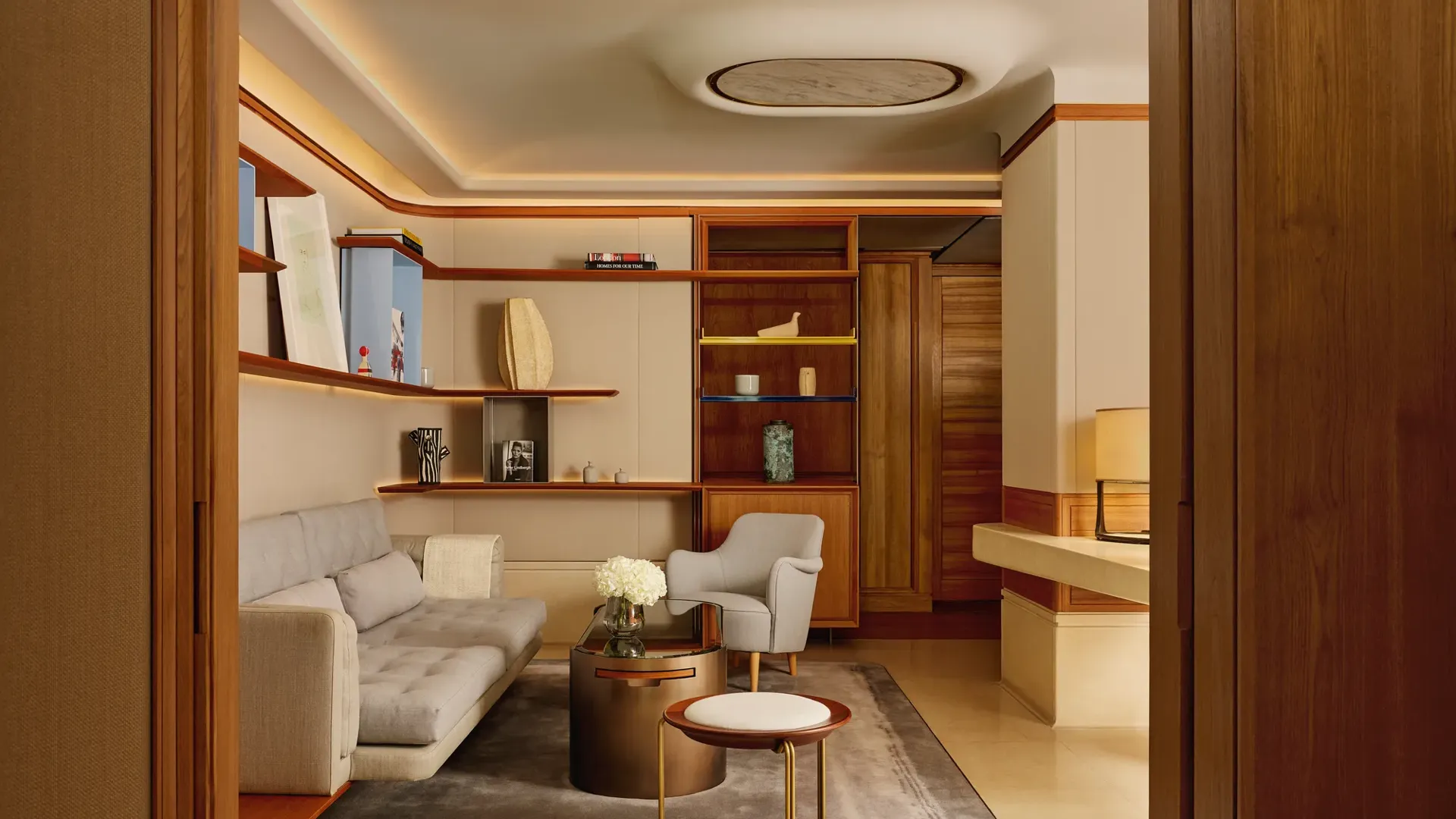 A stylish sitting room with a pale grey sofa, armchair, and round coffee table, framed by wooden shelving displaying books and decorative pieces