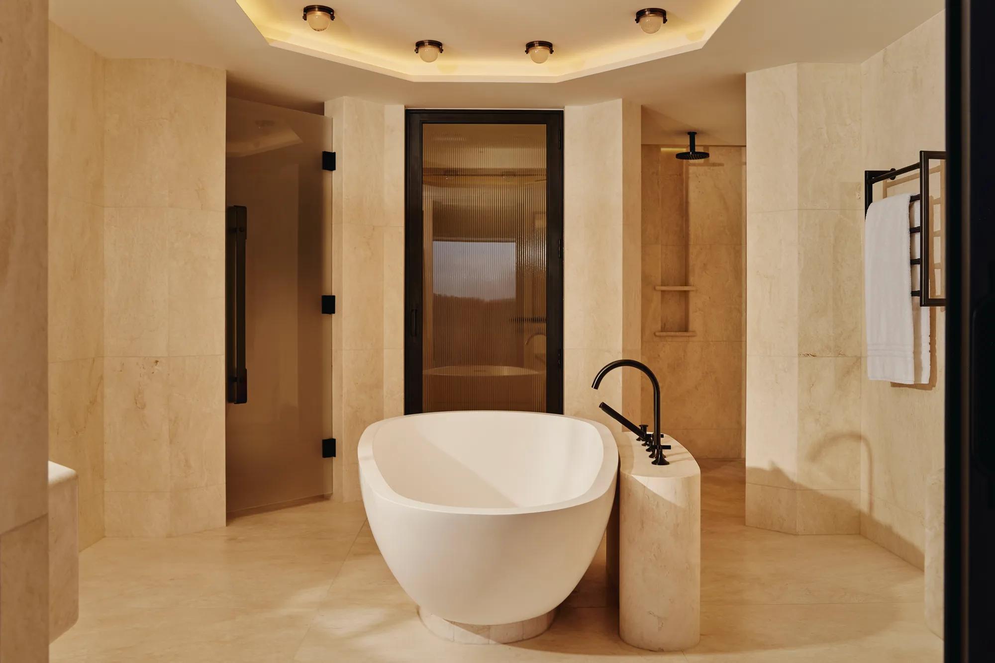 Freestanding oval bathtub in beige marble bathroom with rainfall shower and soft lighting.